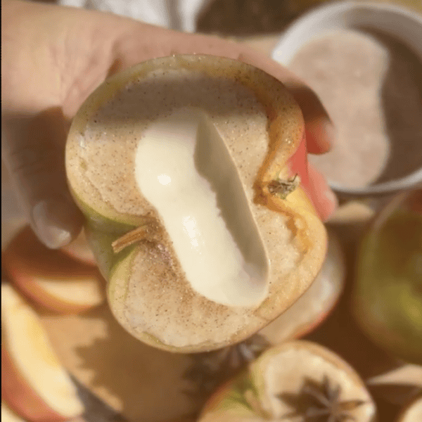 A hand holds a sliced apple with a hollowed-out center filled with cream, dusted with cinnamon.